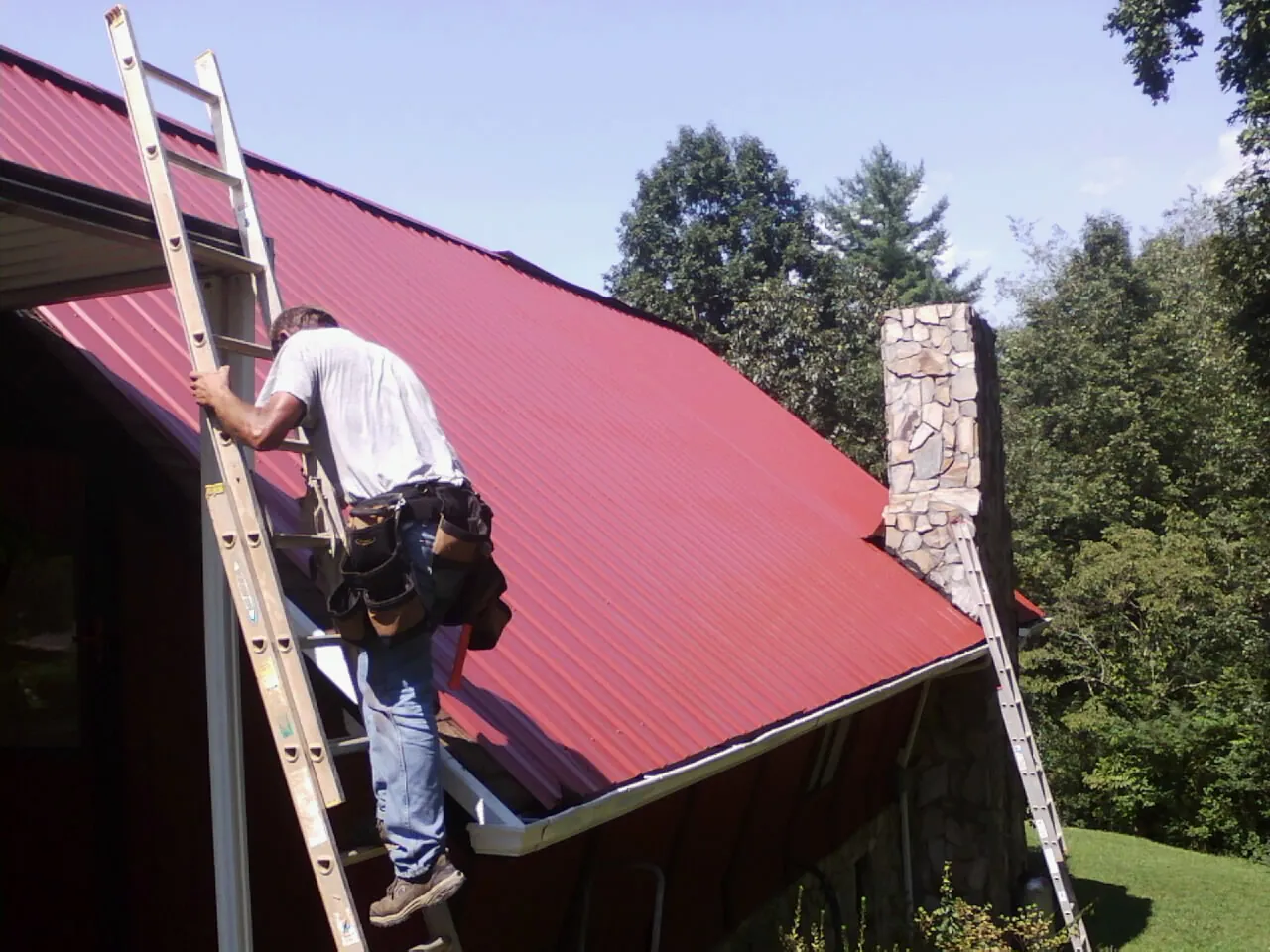 Roofer on a ladder beside a red metal roof and stone chimney — Soffit & Fascia Repair by Roofing Brothers in South Hamilton
